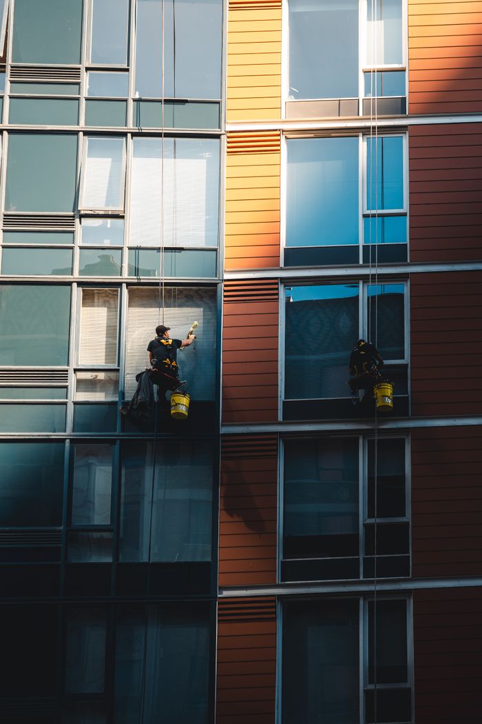 hero-img-01 Two workers hanging to clean windows on a modern skyscraper facade.