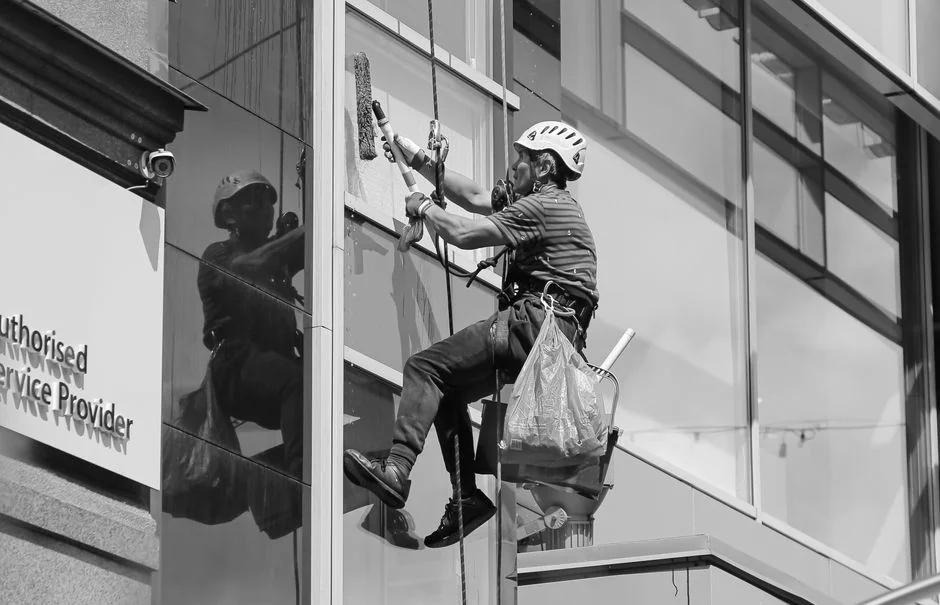 about-01 Two workers clean windows on a modern glass building facade using a suspended platform.